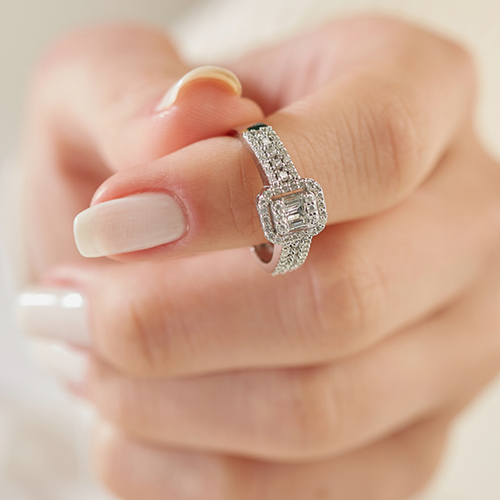 Close-up of a woman's fingers holding a white gold engagement ring featuring an emerald-cut center diamond, a double diamond halo, and a split-shank pavé band.