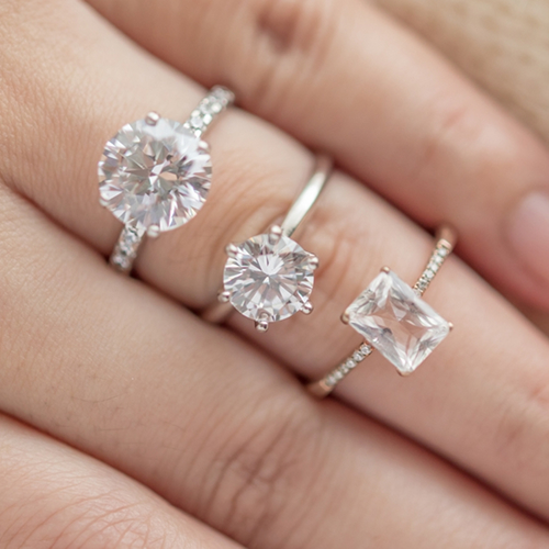 Lifestyle close-up of a hand modeling three different diamond engagement ring styles, including two round brilliant cuts and one radiant-cut diamond on a delicate pavé band.